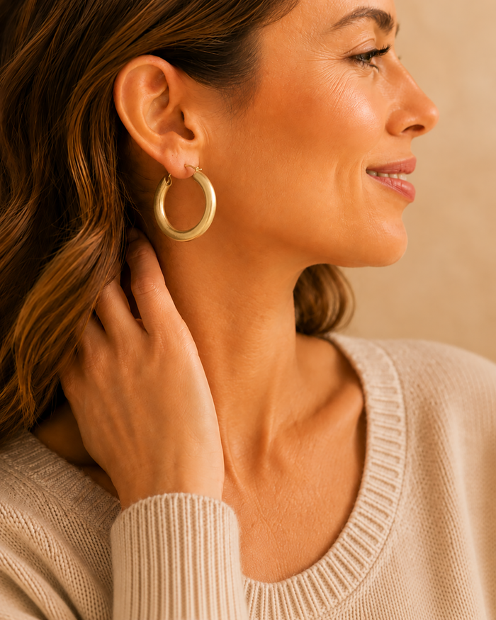 Model wearing large worn-gold tubular hoop earrings with a neutral sweater.