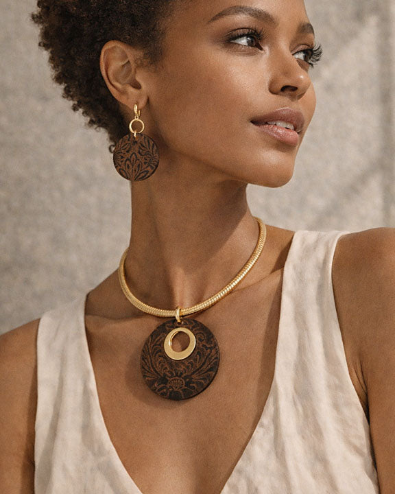 Woman wearing a gold necklace with large wooden pendant and matching earrings against a neutral background