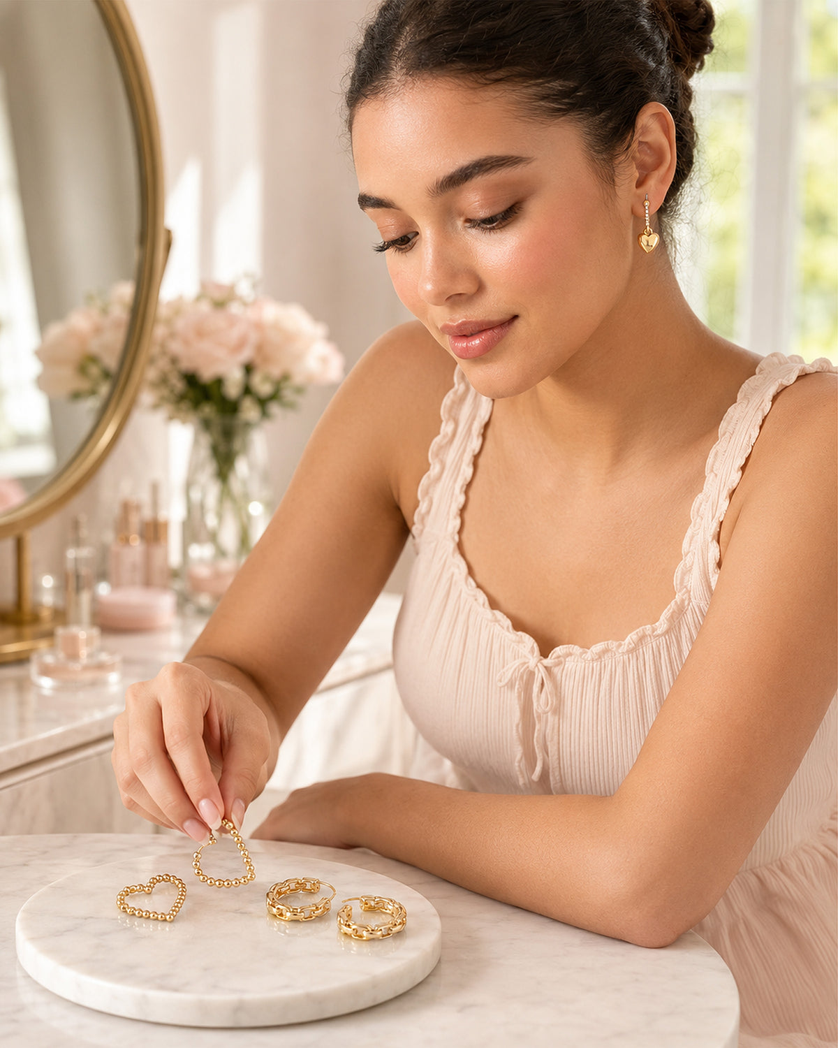 A woman in a light pink pleated top examines gold heart-shaped hoop earrings on a round white marble tray, set in a bright, softly-lit vanity area.