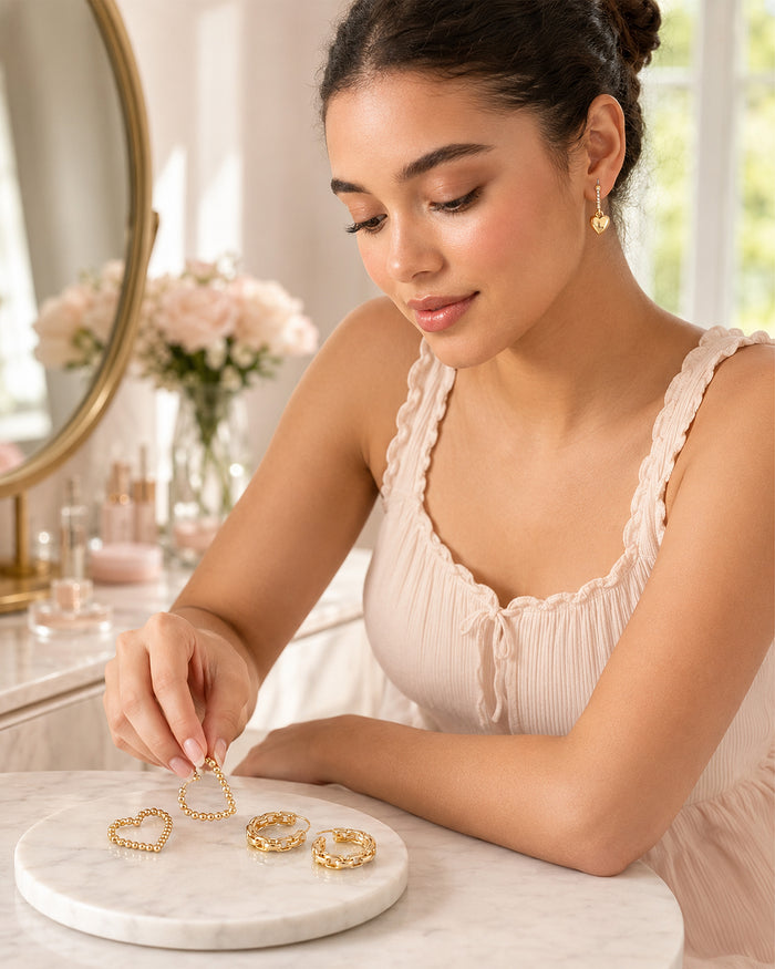 A woman in a light pink pleated top examines gold heart-shaped hoop earrings on a round white marble tray, set in a bright, softly-lit vanity area.
